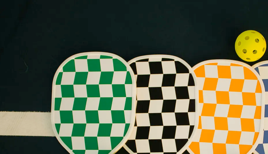 Four checkered pickleball paddles in green, black, orange, and blue with yellow balls on a dark background