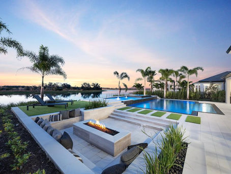 Luxury backyard with blue pool, fire pit, lounge chairs and palm trees beside a calm lake at sunset.
