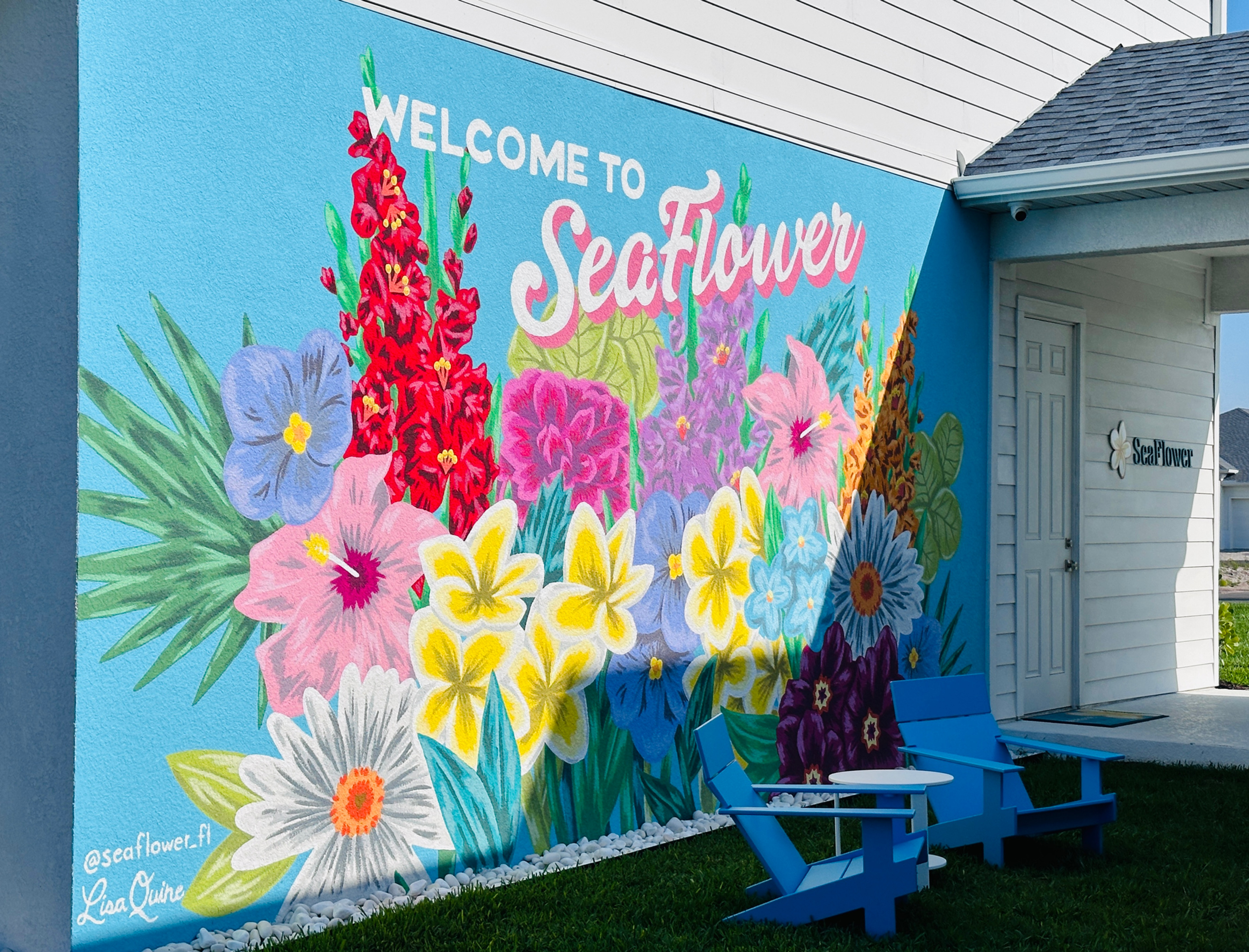 Colorful flower mural on a blue wall with "Welcome to SeaFlower" text. Two blue chairs and a table are on the grass below.