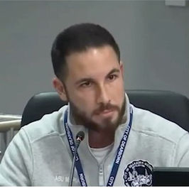 Man with a beard speaking into a microphone at a conference table, wearing a white jacket with a logo. Serious expression, curtain in background.