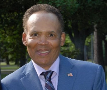 Man in a blue suit and striped tie smiles in a park setting. Trees and grass in the background, creating a cheerful atmosphere.