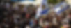 Crowd waving Israeli flags at an outdoor event. The setting is festive with tents in the background and vibrant, sunny lighting.