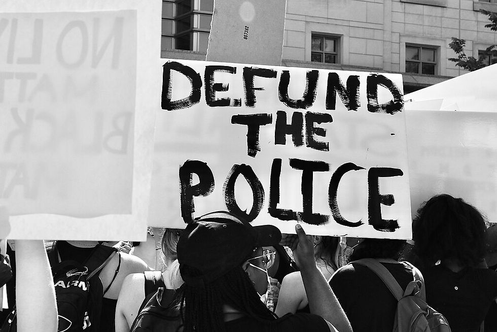 Crowd of people at a protest holding signs, one prominently reads "DEFUND THE POLICE" in bold black letters; urban backdrop.