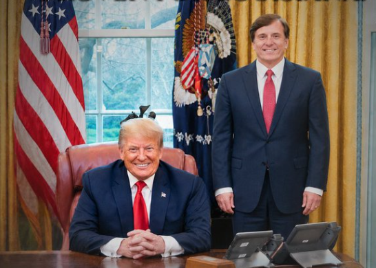 President Trump and John Fleming in suits, one seated at a wooden desk and the other standing, in an office with flags and a window. Both are smiling.
