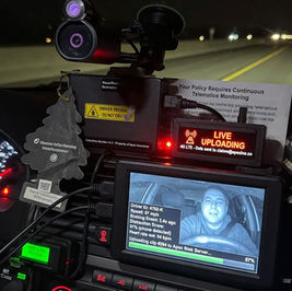 Car dashboard with live telematics monitoring. Screen shows driver's face and data. "LIVE UPLOADING" text in red. Night highway backdrop.