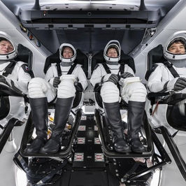 Four astronauts in white space suits sit in seats inside a spacecraft. They appear calm and focused. The interior is sleek and modern.