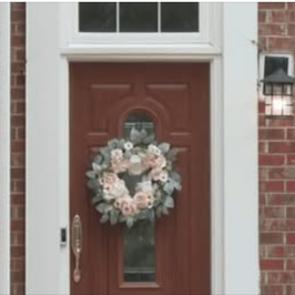 A brown door with a floral wreath, set in a red brick wall. A black lantern is mounted near the door, creating a welcoming atmosphere.