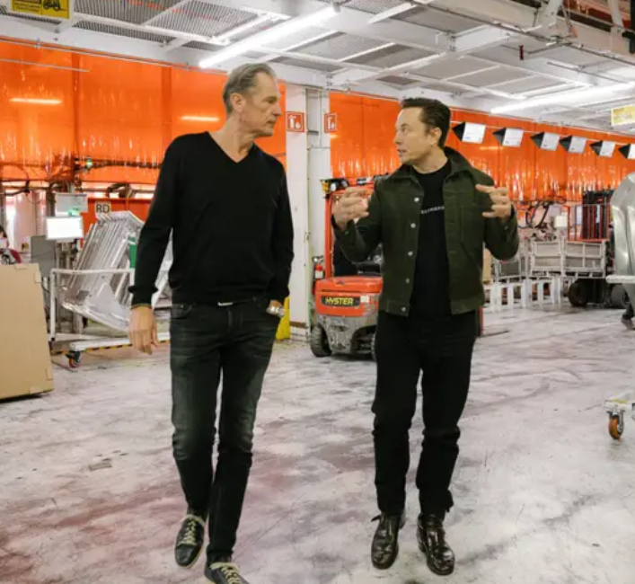 Two men walk and talk in an industrial warehouse with orange barriers; one gestures while explaining.