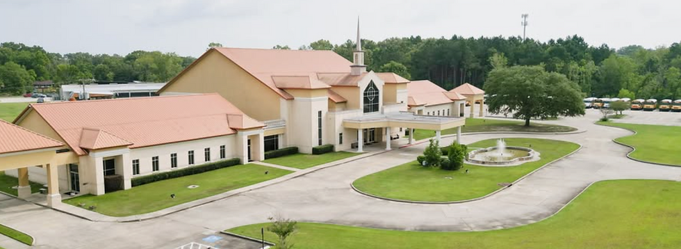 Large beige church with red roofs and steeple, circular fountain, empty drive, and school buses near trees on a cloudy day.