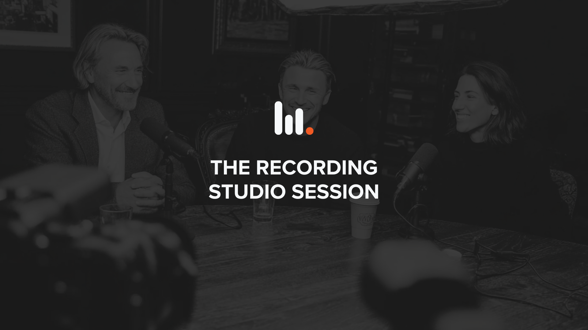 Black-and-white image of three people recording a podcast at a table with microphones, cameras in the foreground, and studio lighting above in a podcast studio setting.