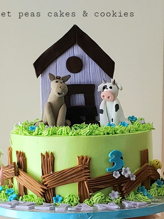 Farm-themed birthday cake with green icing, fondant animals (pig, cow, dog, donkey), wooden fence, barn centerpiece, blue number 3, and piped grass and flowers.