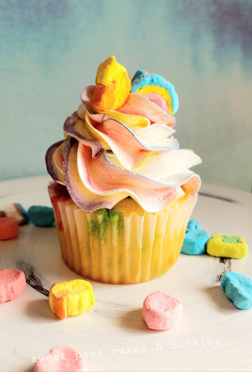 Rainbow swirl cupcake with pastel buttercream and colorful cereal marshmallows on top, surrounded by extra marshmallows.