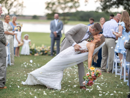 Bride and groom dip kiss at outdoor ceremony at The Vincent Homestead with lake in background and white rose petals lining the aisle — Central Texas wedding venue