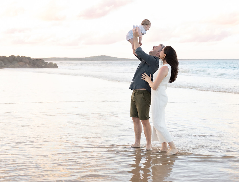 Mum and dad holding their baby in the air at Noosa Main Beach during an outdoor family photography session.