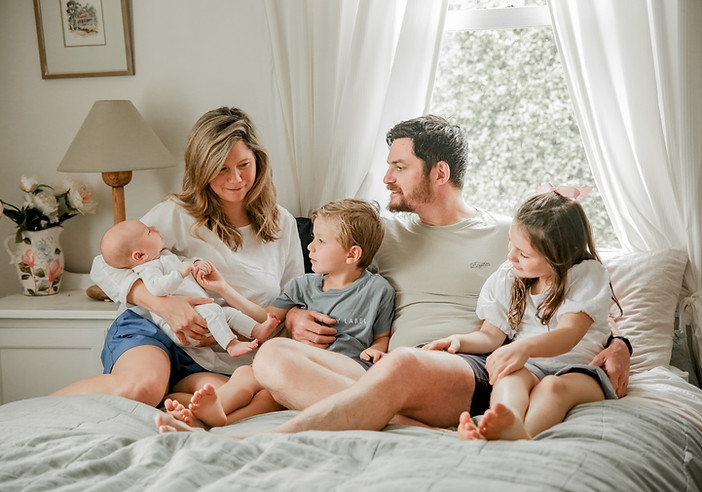 Mum, dad, and two older children sitting on a bed looking at their newborn baby during an in-home Noosa family photo session