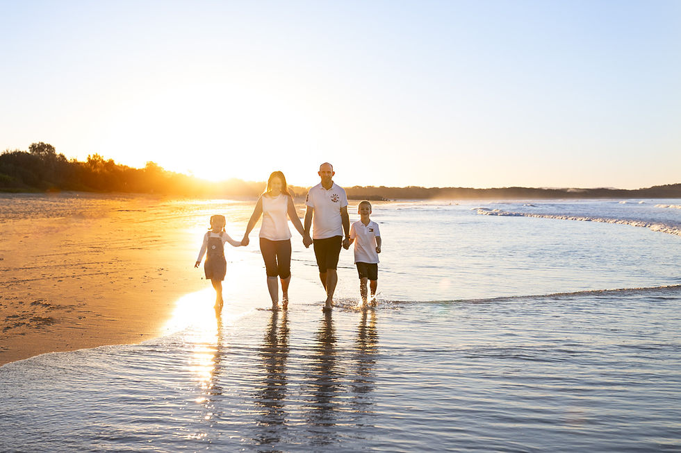 Silhouette of a family of four walking hand-in-hand through the shallow ocean water, beautifully backlit by the golden setting sun during a Noosa beach photography session.