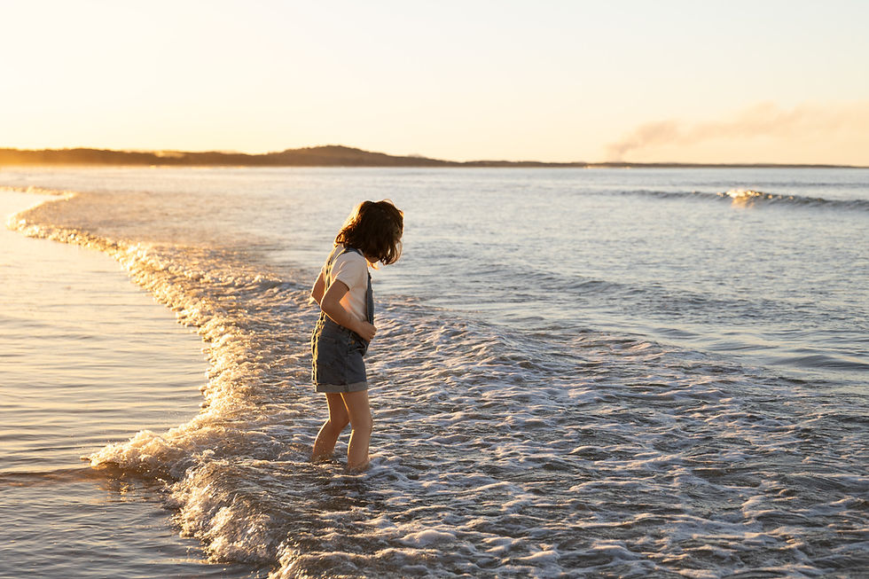 Candid photo of a young girl walking through the shallow ocean water, silhouetted against a vibrant golden hour sunset on a Noosa beach.