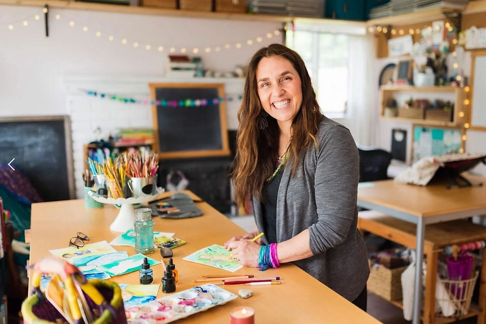 Photo of Illustrator Faith Pray standing at a table in her studio, with paints and brushes near her.