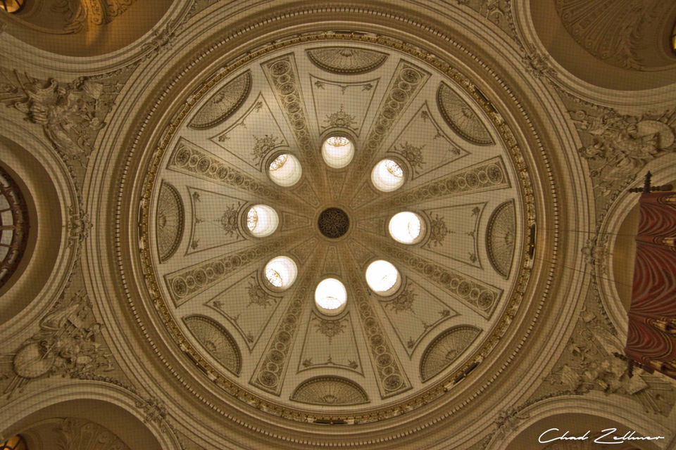 Looking upward at an ornate circular ceiling with carved details and bright round windows.