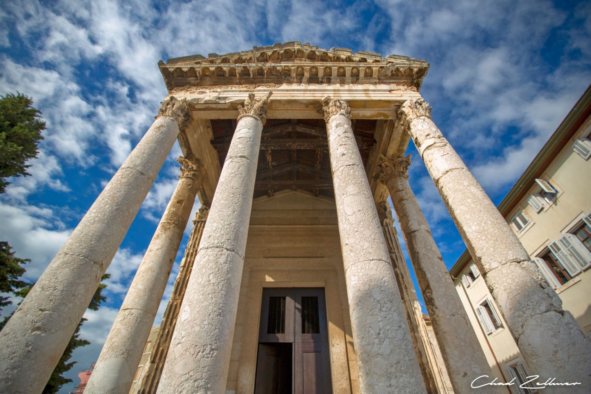 Tall weathered stone columns support an ornate classical pediment beneath a blue sky with streaked white clouds.