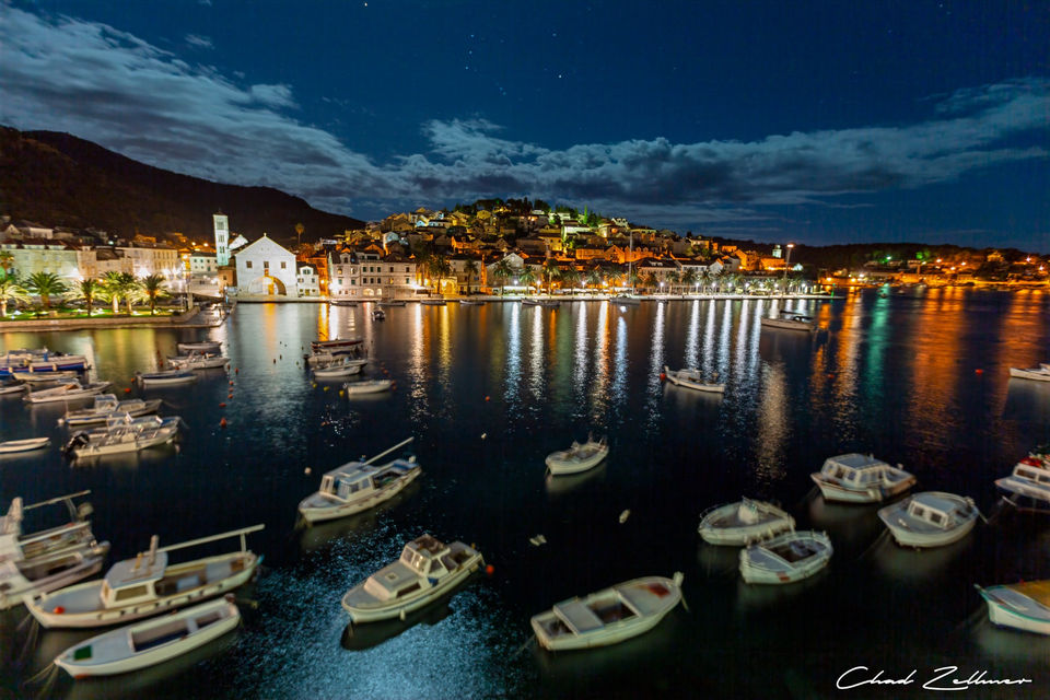 Small boats float in a dark harbor beneath illuminated waterfront buildings, hillside homes and a cloud streaked night sky.