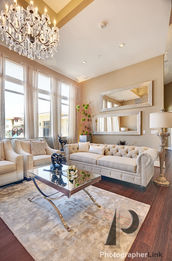 Living room with crystal chandelier, tufted beige sofa and armchairs, glass coffee table, tall windows, plush rug, and decorative mirrors on neutral walls.