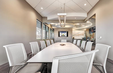 Contemporary conference room featuring a long white table, ergonomic mesh chairs, glass walls, ceiling lights, and wall-mounted digital display
