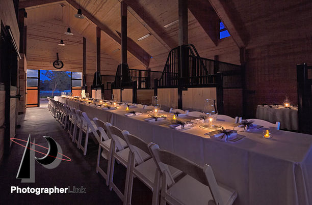 Long banquet table with white chairs set inside a vaulted wooden barn, softly illuminated by candles at dusk.