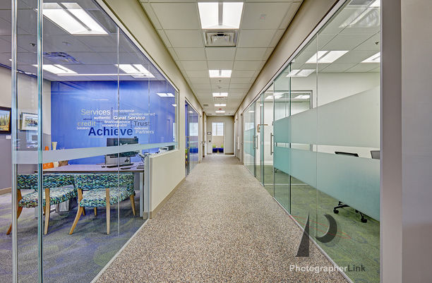 Office hallway with glass walls for meeting rooms, blue branded wall with motivational words, patterned lounge chairs, workstation, neutral carpet, and ceiling lights in a modern workplace.