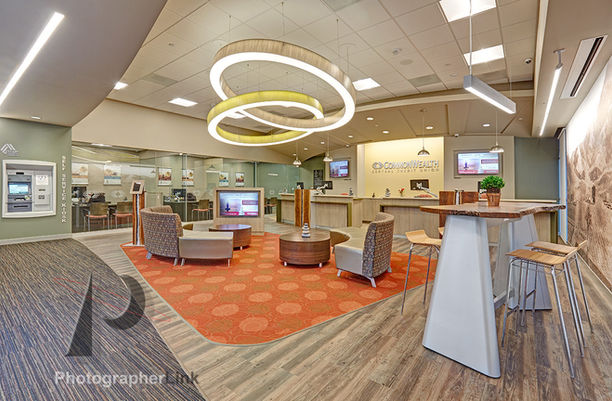 Modern lobby area at Commonwealth Central Credit Union with lounge seating, ATM kiosk, wood reception desk, circular pendant lighting, and digital monitors.