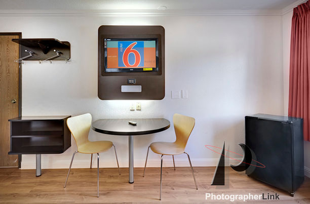 Motel 6 room with wall-mounted TV, small table and chairs, mini fridge, coat rack, wood flooring, and light walls.
