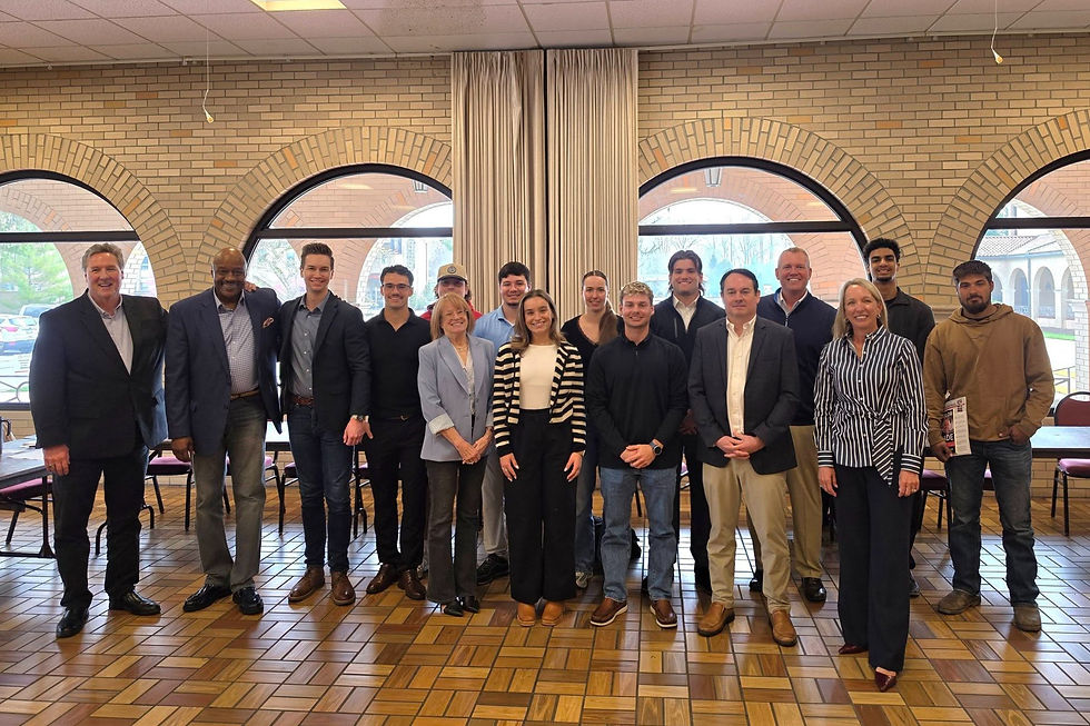 Group photo of Taylor Automotive Family speakers and Lourdes University attendees standing together inside a campus room with arched windows.