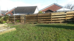 A back garden with a wooden fence, stacks of lumber, and neatly cut grass