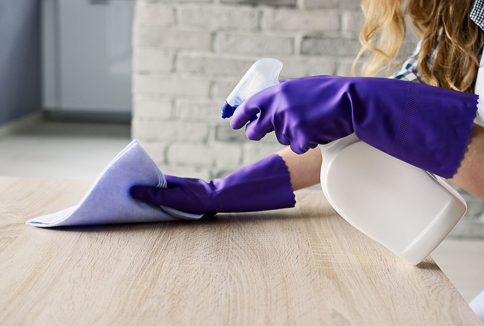 A person wearing purple gloves cleans a wooden table with a spray bottle and cloth