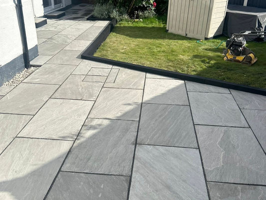 A sunlit patio with large gray tiles leads to a small grassy area. In the corner, there’s a beige storage shed