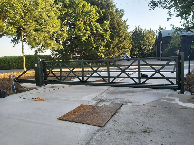 Green metal gate across rural driveway, with trees and barn under sunny sky