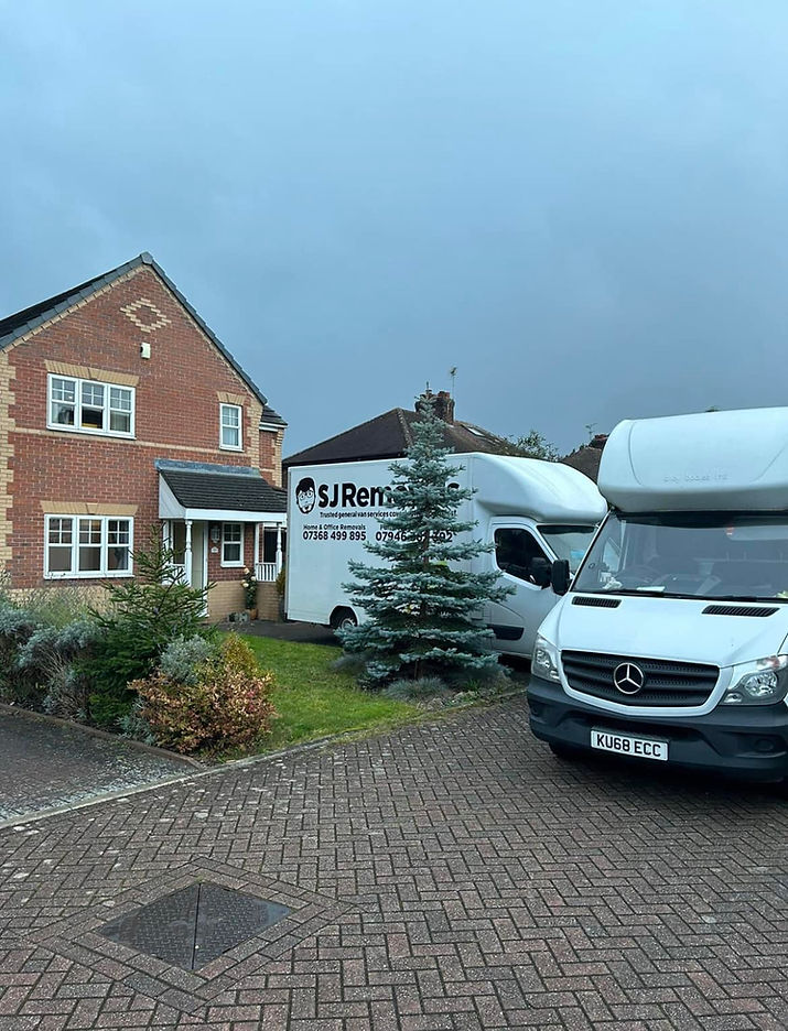 A suburban brick house with a garden is seen with two white moving vans parked in the driveway