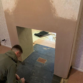 A person in a green jacket works on laying dark tiles on a floor near a wall