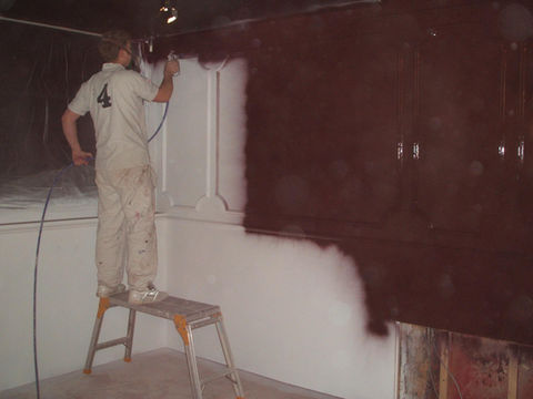 A person stands on a ladder, spray-painting a wall dark brown