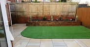 A tidy backyard features a small artificial grass area bordered by beige tiles