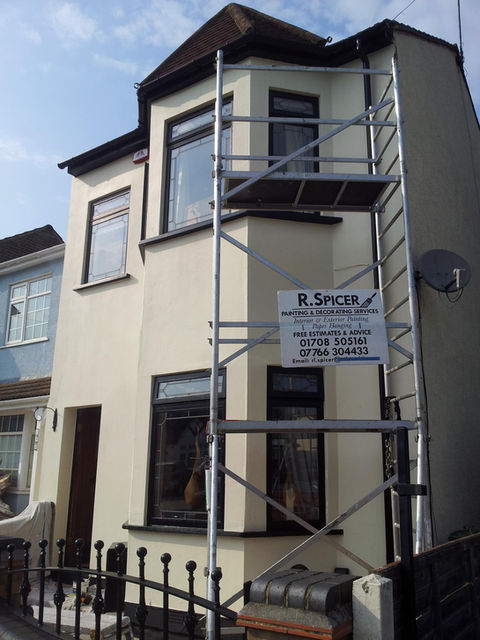 A two-story house with pale exterior walls and a dark roof, featuring scaffolding on the front