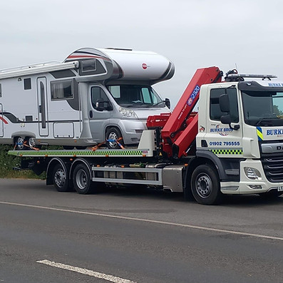 A truck, equipped with a red crane, is transporting a large white motorhome on a roadway
