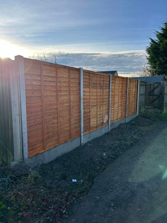Wooden fence with vertical slats bordered by concrete posts runs along a narrow path