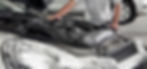 A person in a white shirt inspects a car engine under an open hood in a garage