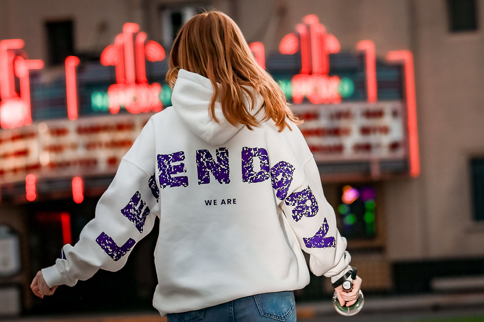 Lifestyle photo of a person wearing a white hoodie with bold purple text on the back, standing in front of colorful neon lights