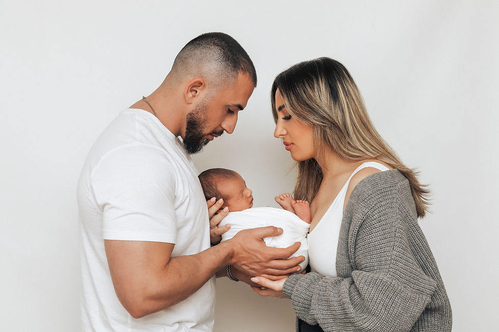 A couple lovingly gazes at their swaddled newborn between them in a serene setting. Both wear casual attire; the background is plain white.