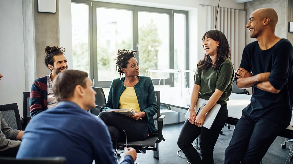 A diverse group of people sits and stands in an office, engaged in lively conversation and laughing. Bright, modern setting with large windows.
