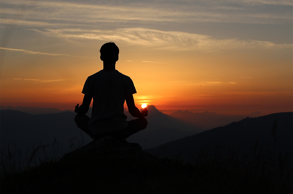 Silhouette of a person meditating on a hill at sunset. The sky is a gradient of orange and yellow with mountains in the background. Peaceful mood.