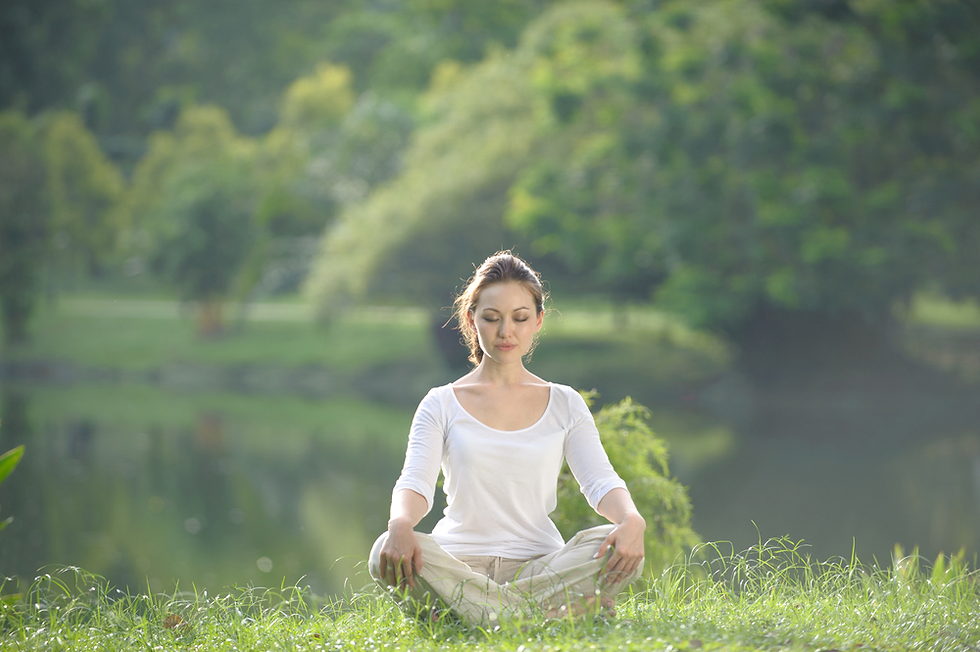 Woman meditating in a green park by a pond, wearing a white shirt and beige pants. The serene setting includes lush trees and grass.