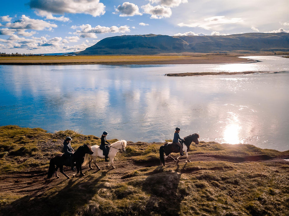 Three riders on horses travel along a grassy riverbank under a bright cloudy sky, with mountains reflected in calm water.